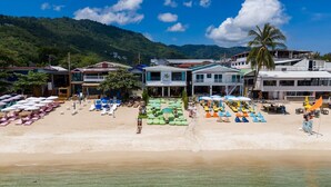 Un hotel frente a la playa con varias habitaciones, una playa de arena y montañas al fondo.