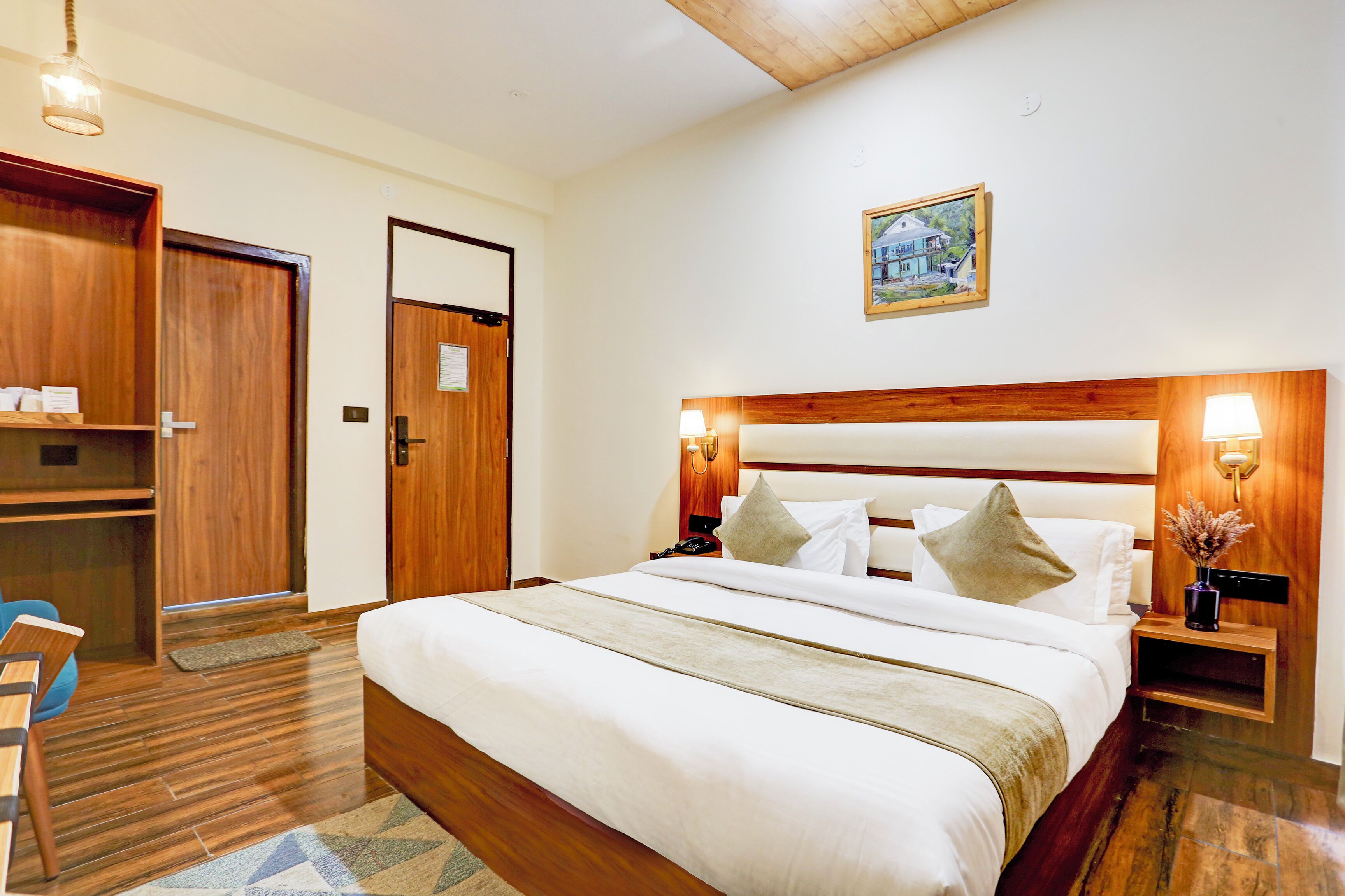 A hotel room with a large bed, wooden headboard, bedside lamps, a wooden wardrobe, and a framed picture on the wall.