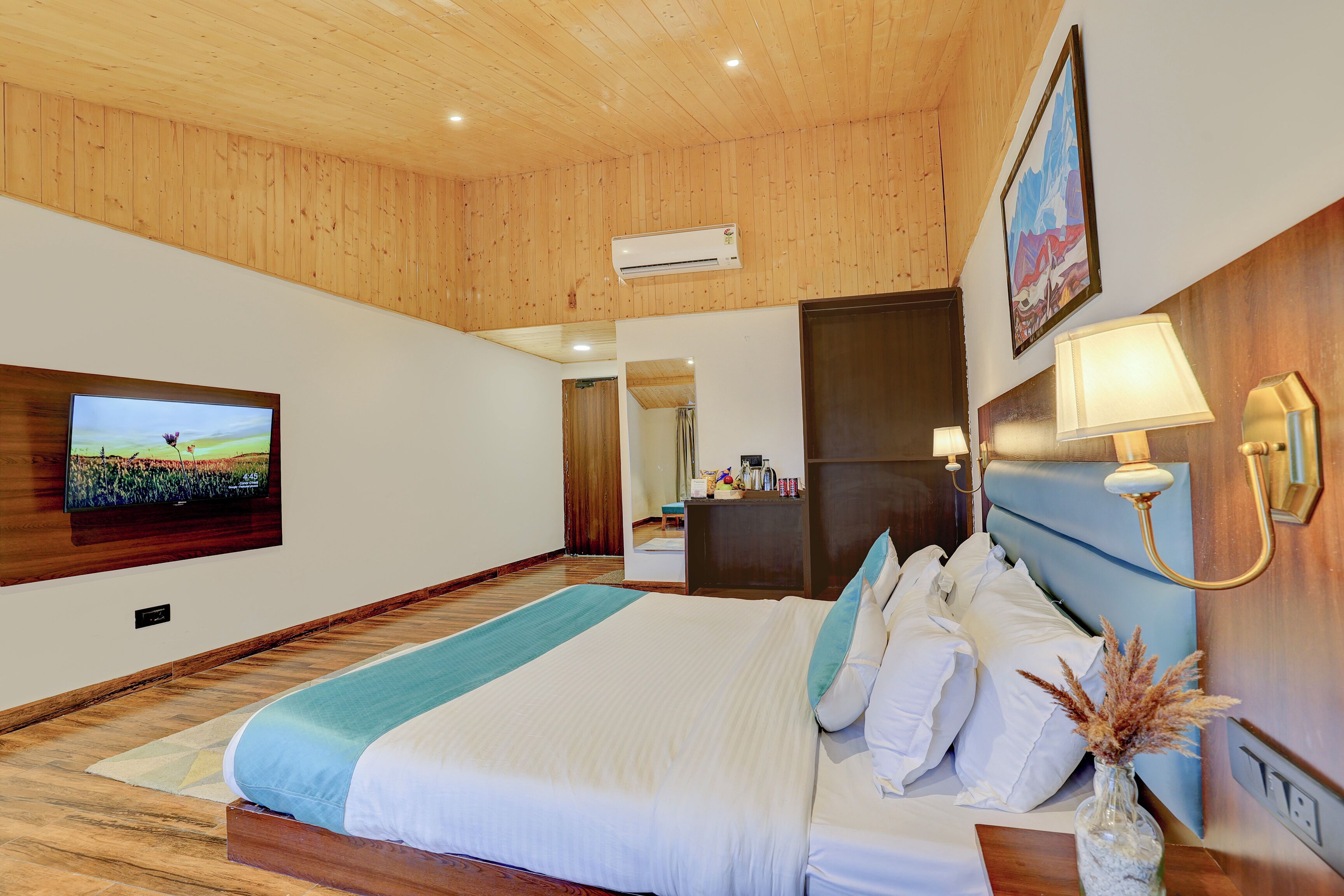 A spacious bedroom with a large bed, wooden ceiling, a flat-screen TV, a bedside table with a lamp, and a mirror.