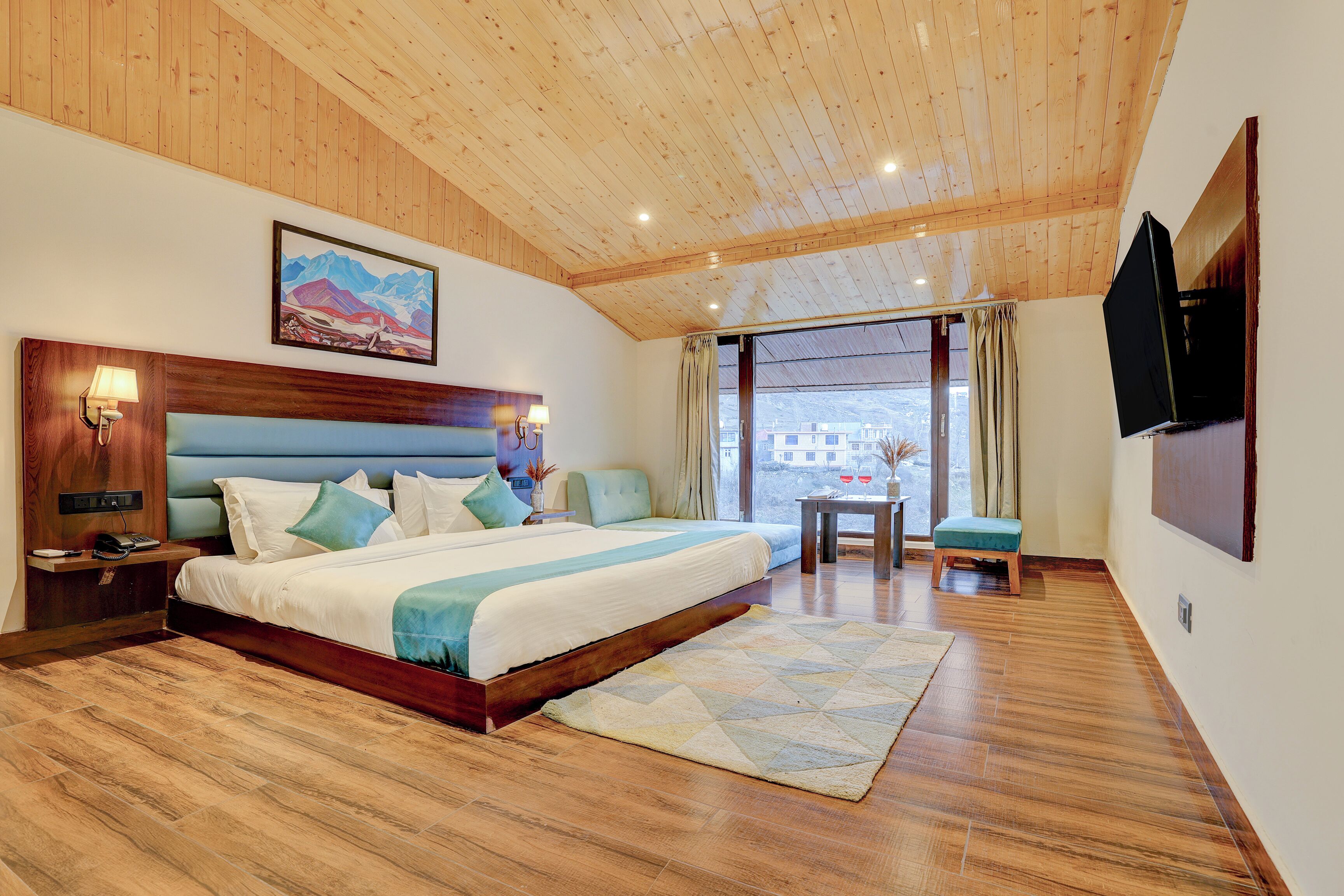 A bedroom with a wooden ceiling, a large bed with a headboard, a bedside table with a lamp, a small table with a vase, and a window with curtains.