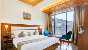 A hotel room with a large bed, wooden headboard, two bedside tables with lamps, a small table with chairs, and a large window with a view of buildings. - Utpala House By Lifestyle (Lahul)