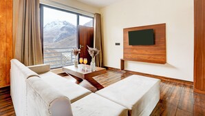 A modern hotel room with a large window, wooden flooring, a sofa, a table with drinks, and a TV mounted on the wall. - Utpala House By Lifestyle (Lahul)