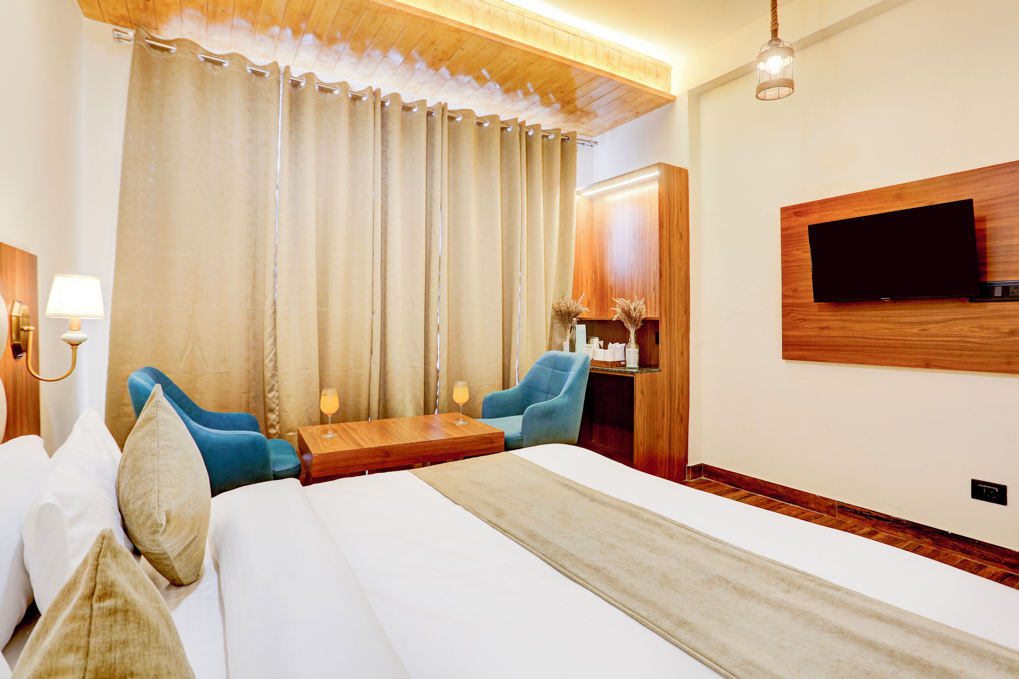 A hotel room with a bed, two chairs, a small table, a TV mounted on the wall, and a wooden ceiling.
