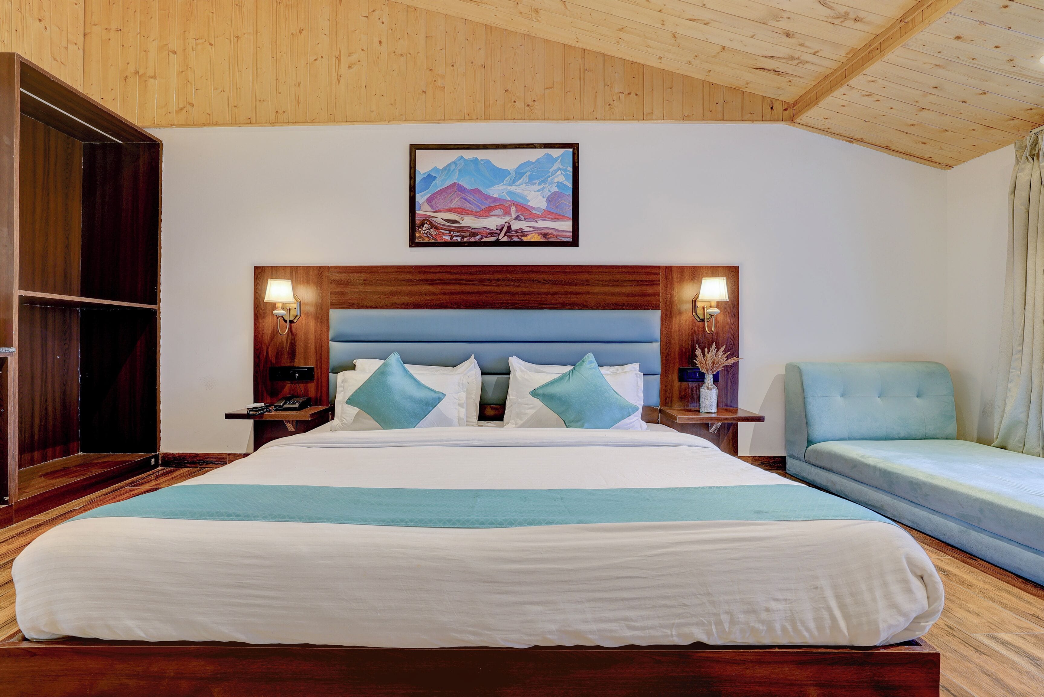 A large bed with a wooden headboard, two bedside tables with lamps, a light blue sofa, and a painting of mountains on the wall.