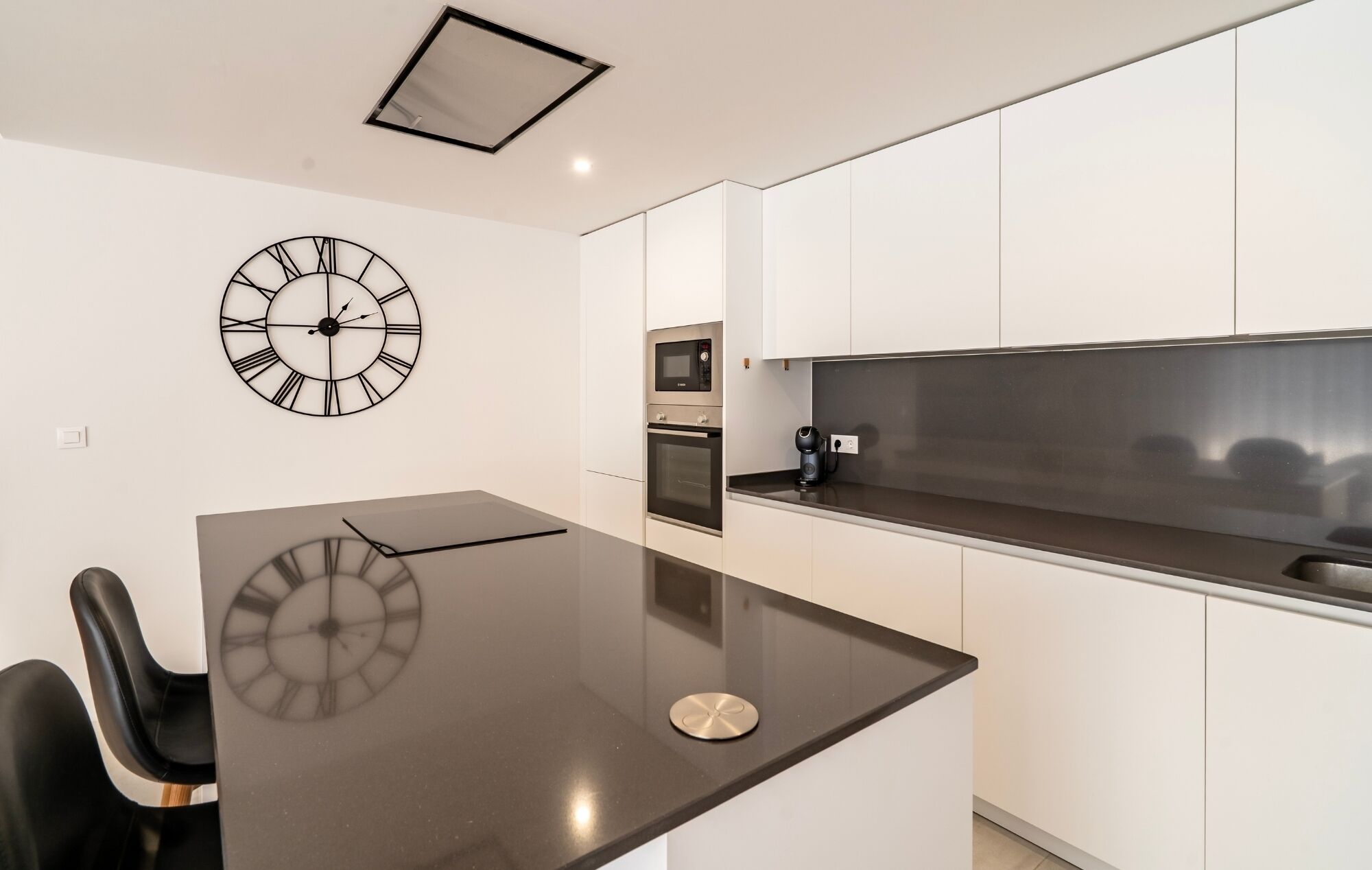A modern kitchen with white cabinets, a black countertop, a stainless steel range hood, and a wall clock.