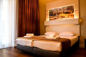 A hotel room with a large bed, a headboard with a cityscape artwork, and a window with curtains.