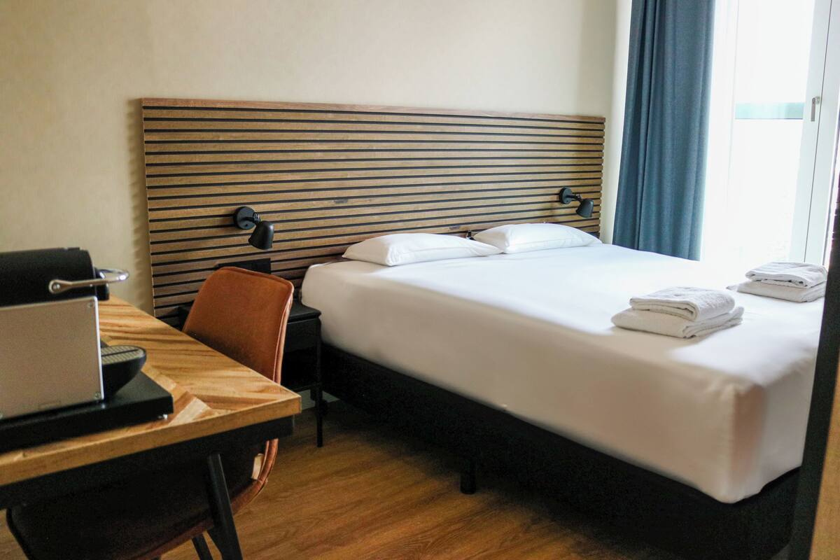 A hotel room with a bed, a desk with a laptop, a chair, and a window with curtains.