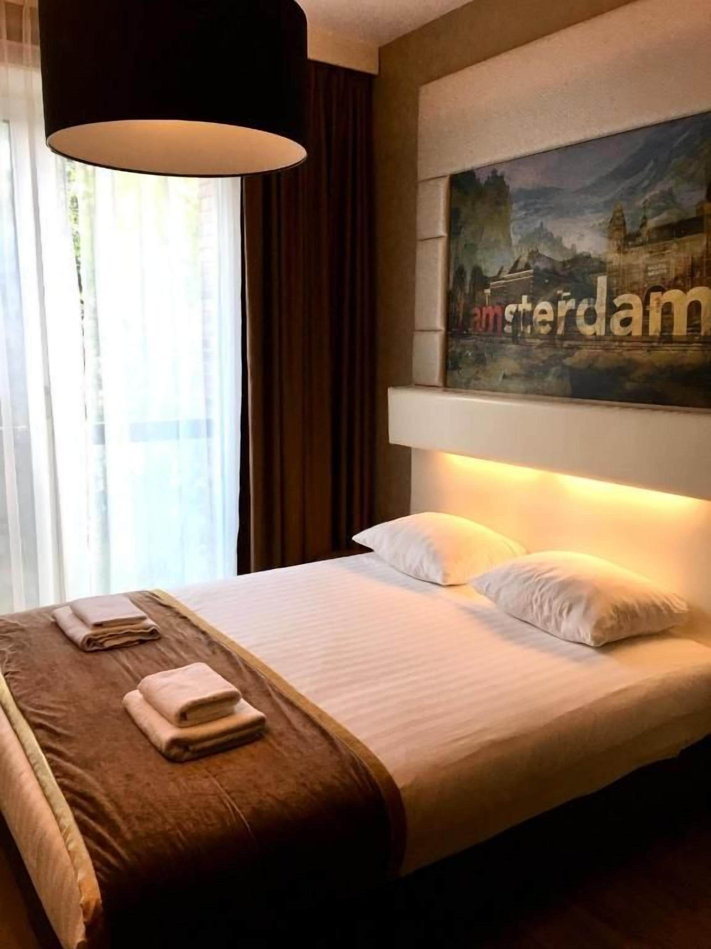 Hotellihuone, jossa on sänky, kaupunkimaisemaa esittävä taulu ja ikkuna, jossa on verhot.