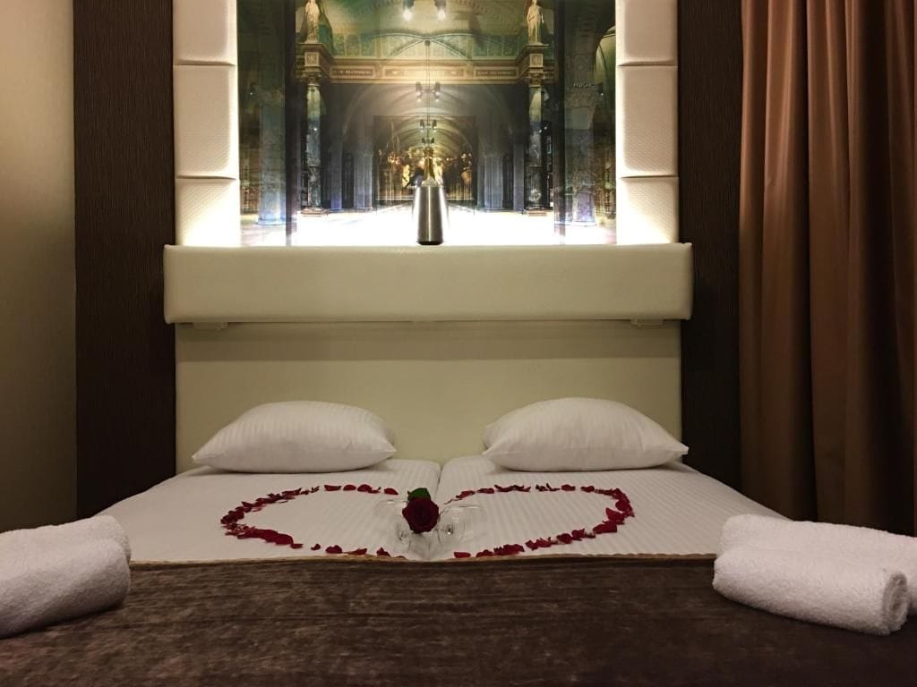 Habitació d'hotel amb un llit al centre sota un gran finestral, un arranjament de pètals de rosa en forma de cor al llit, i una estàtua visible a través de la finestra.