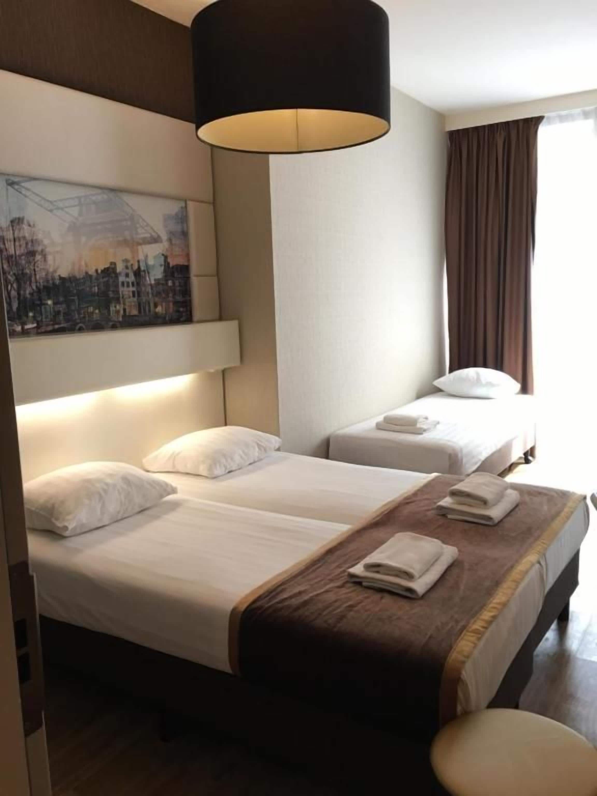 Een hotelkamer met twee bedden, een groot raam met gordijnen, een schilderij aan de muur en een hanglamp.