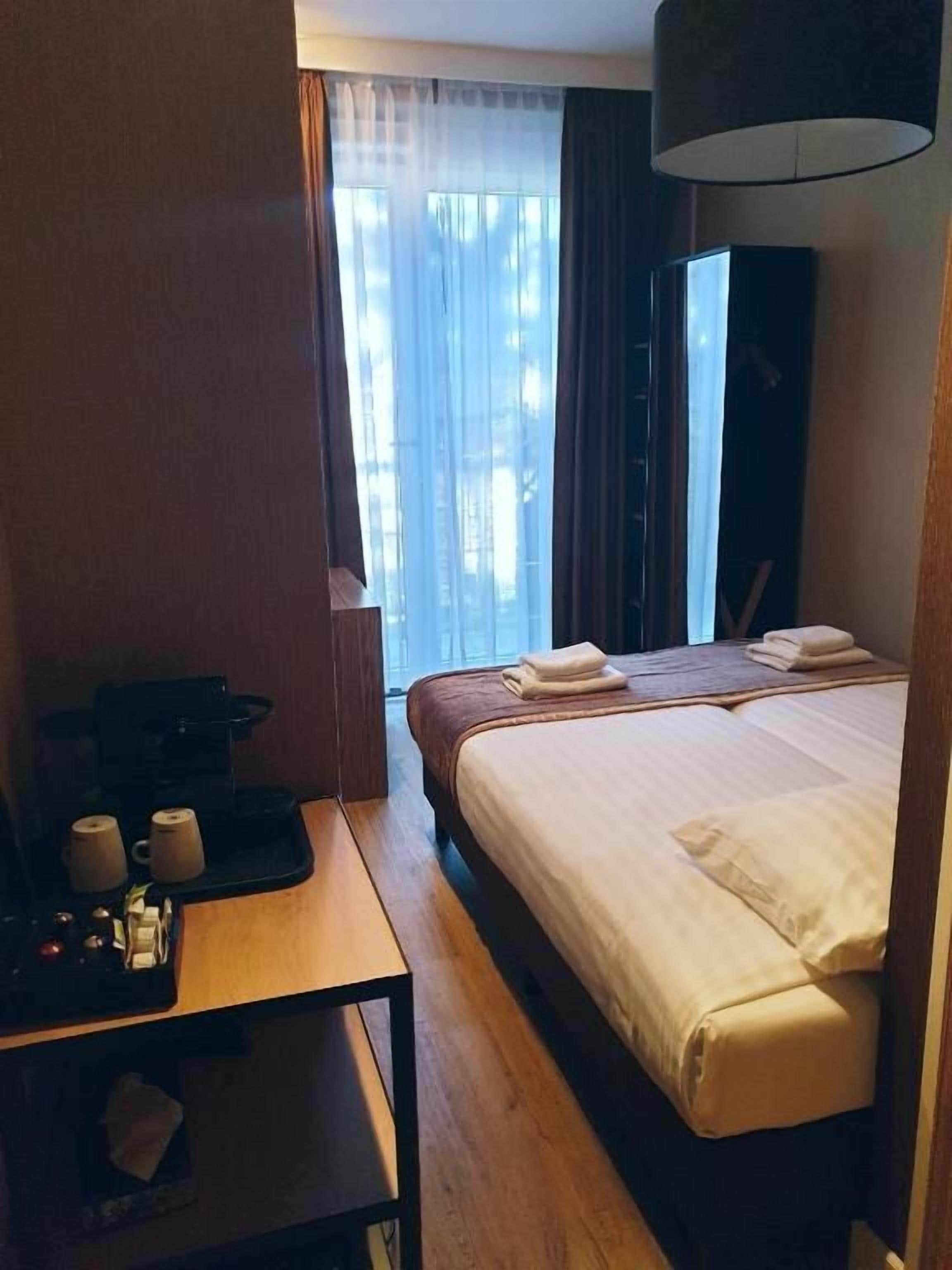 A hotel room with a bed, a table with a coffee maker, and a window with curtains.