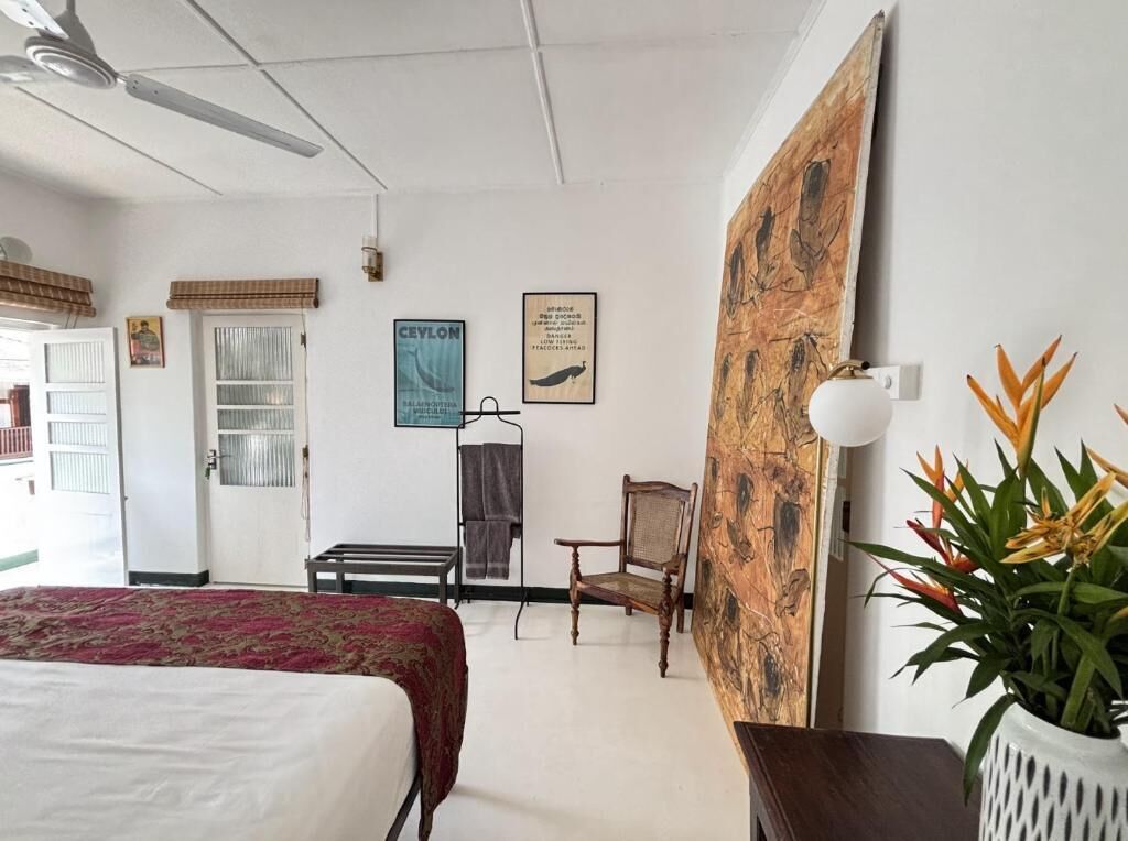 A hotel room with a bed, a chair, a wall with artwork, a ceiling fan, and a potted plant.