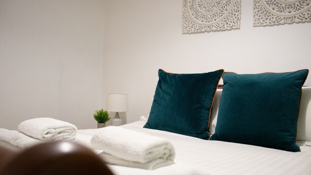 A neatly made bed with white linens and teal pillows, a small bedside plant, and a white wall with decorative wall art.