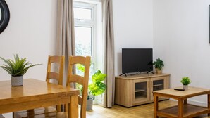 A living room with a wooden dining table, chairs, a TV on a wooden stand, and potted plants.