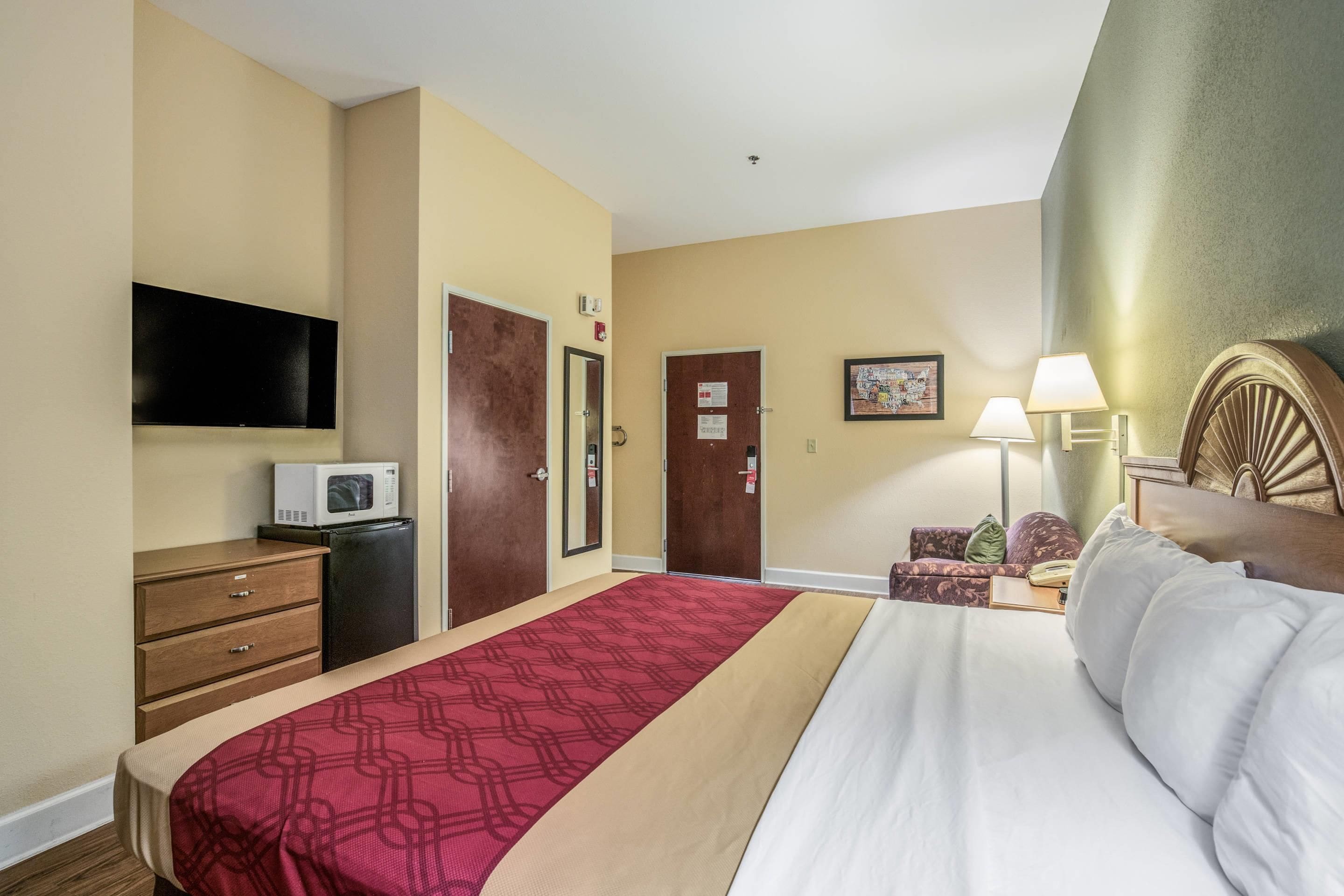 A hotel room with a bed, a television, a microwave, a desk with a lamp, and a door.