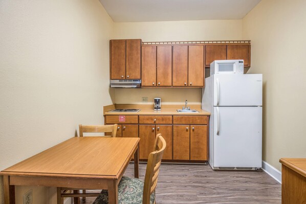 A compact kitchen with wooden cabinets, a white refrigerator, a microwave, and a dining table with chairs. - Econo Lodge Valdosta (Valdosta)