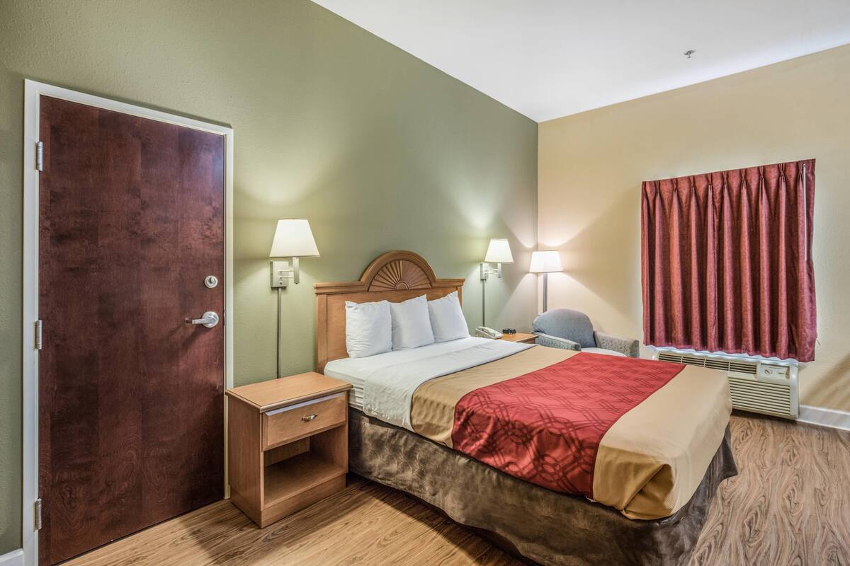 Ein Hotelzimmer mit einem hölzernen Bett, Nachttischen, Lampen, einem Fenster mit Vorhängen und einer Tür.