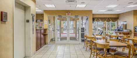 Un vestíbulo de hotel con mostrador de recepción, puerta de entrada de vidrio y un área de comedor con mesas y sillas.