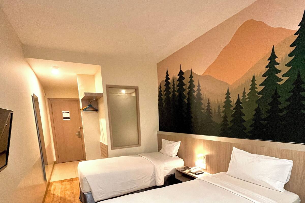 A hotel room with two beds, a wooden floor, a painting of trees and mountains on the wall, and a door leading to a hallway.