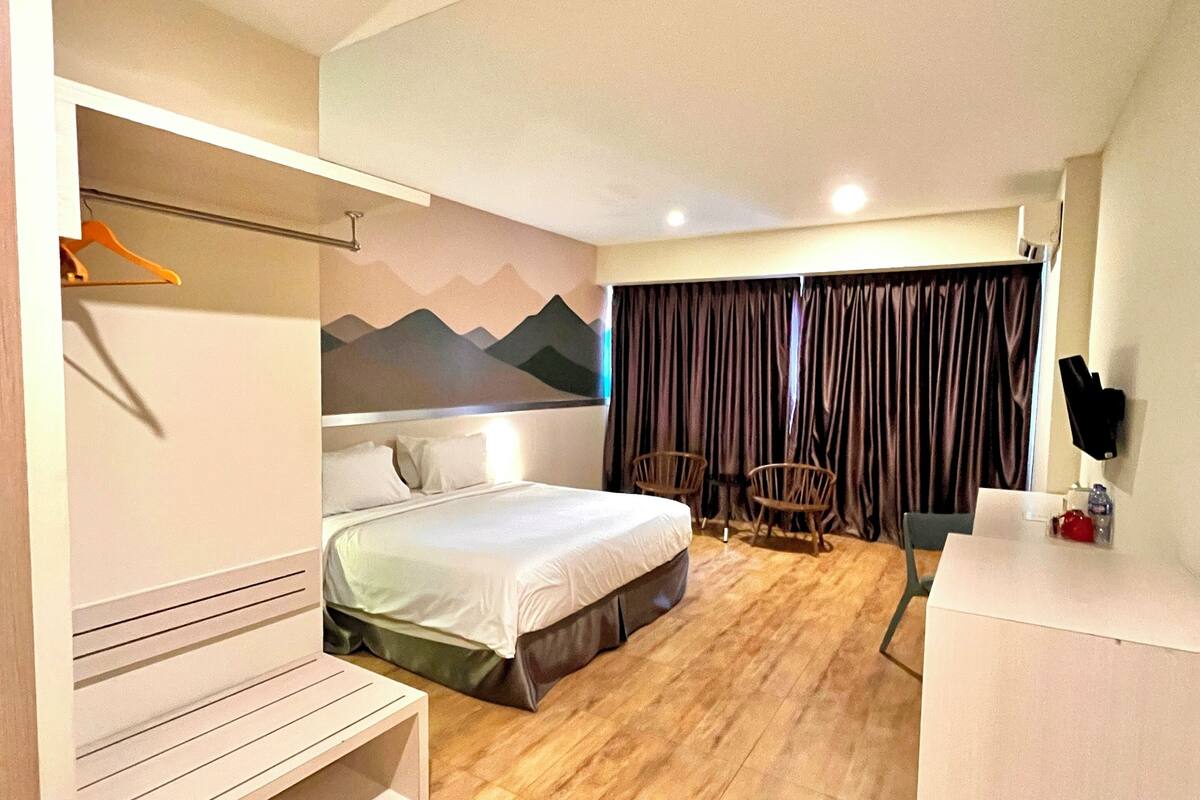 A hotel room with a bed, a desk, a chair, a wardrobe, and a wall with a mountain mural.