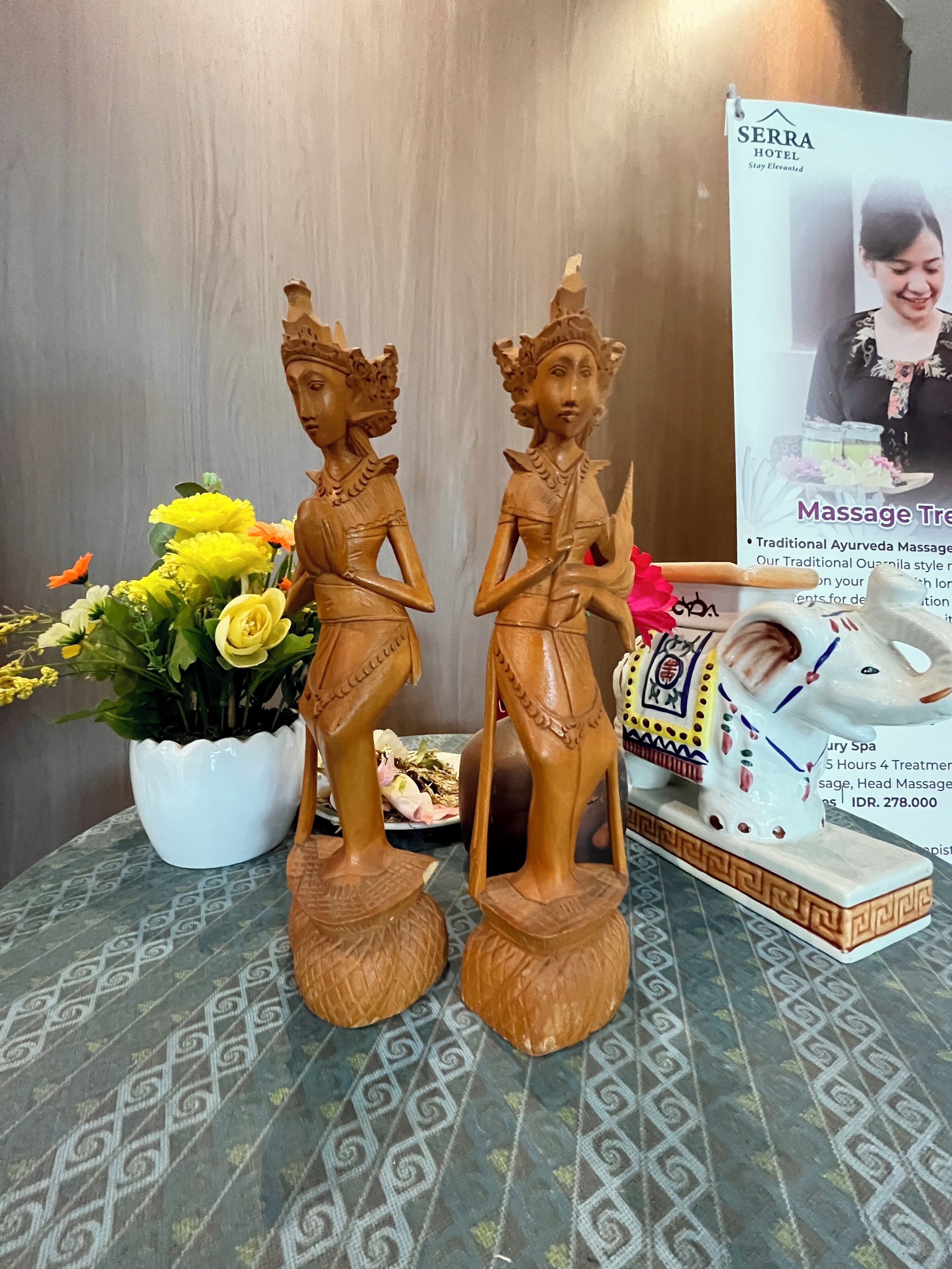 Une statue en bois représentant une figure humaine, une plante en pot ornée de fleurs jaunes et une affiche publicitaire pour un soin de massage.