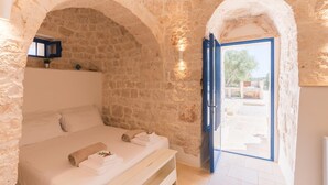 A bedroom with stone walls, a bed with white linens, a bench, and a view through an open door. - Trulli Scacciapensieri con Piscina (Ceglie Messapica)