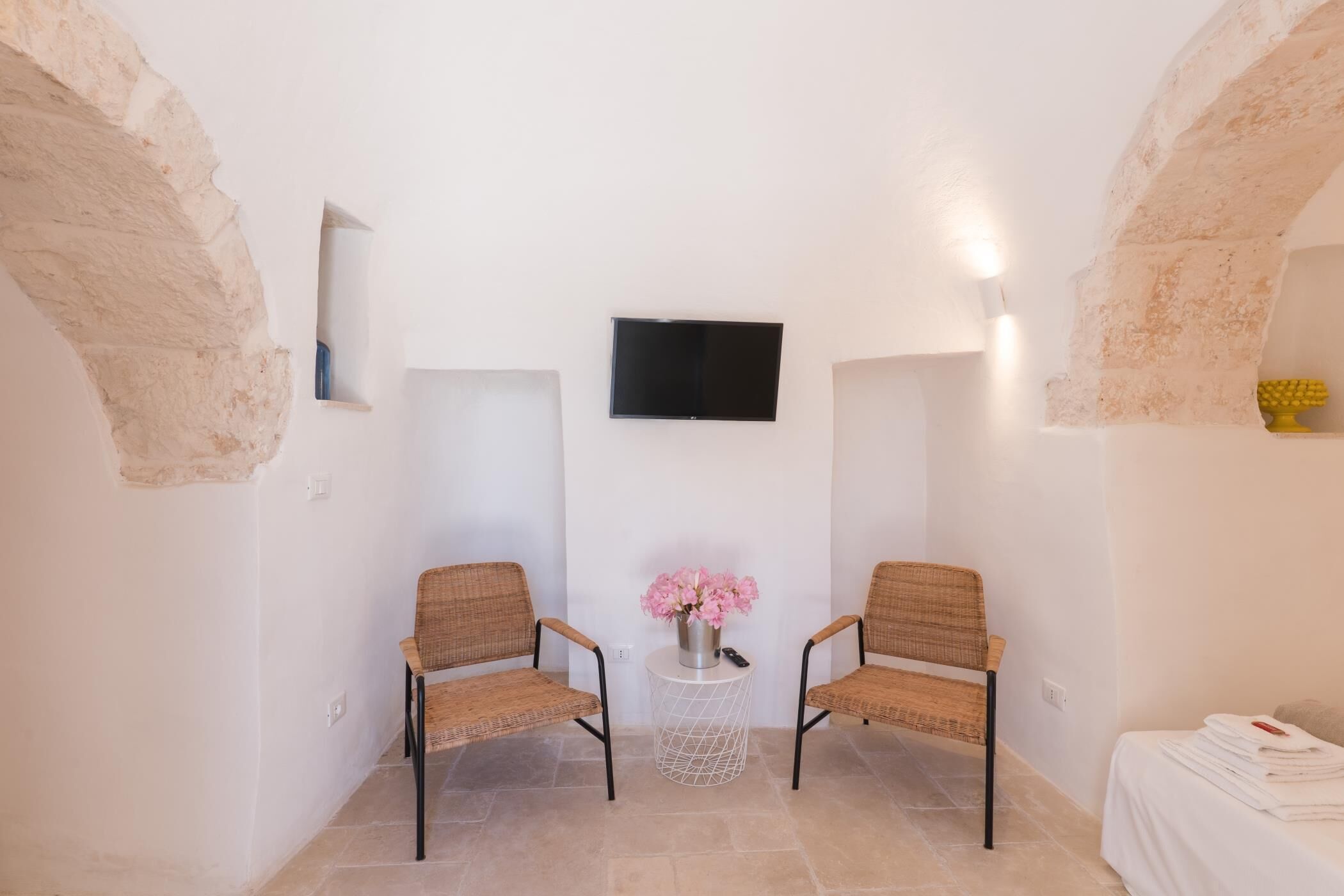 Ein kleiner, gemütlicher Raum mit zwei Korbstühlen, einem Tisch mit Blumenvase, einem flachbildfernseher an der Wand und einem Bett mit ordentlich gefalteten Handtüchern.