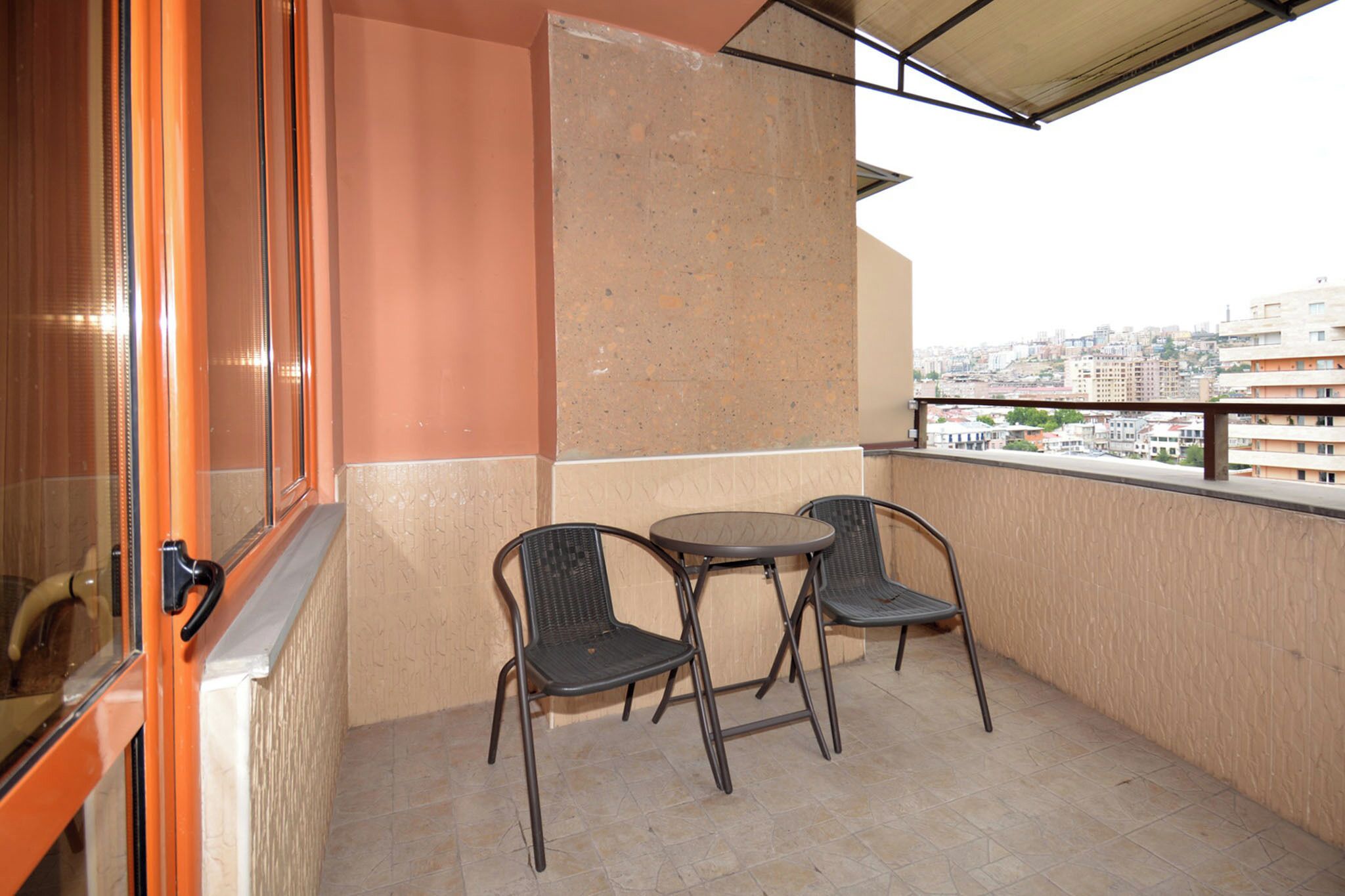 Een balkon met twee stoelen en een tafel, met uitzicht op een stadsgezicht, en een deur met een hendel.