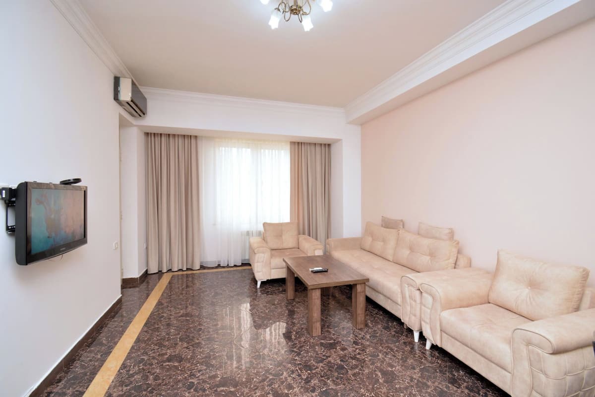 Un vaste salon comprenant un grand canapé beige, un canapé plus petit et une table basse en bois. Un téléviseur à écran plat est fixé au mur, et le sol est recouvert de marbre avec une bande jaune.