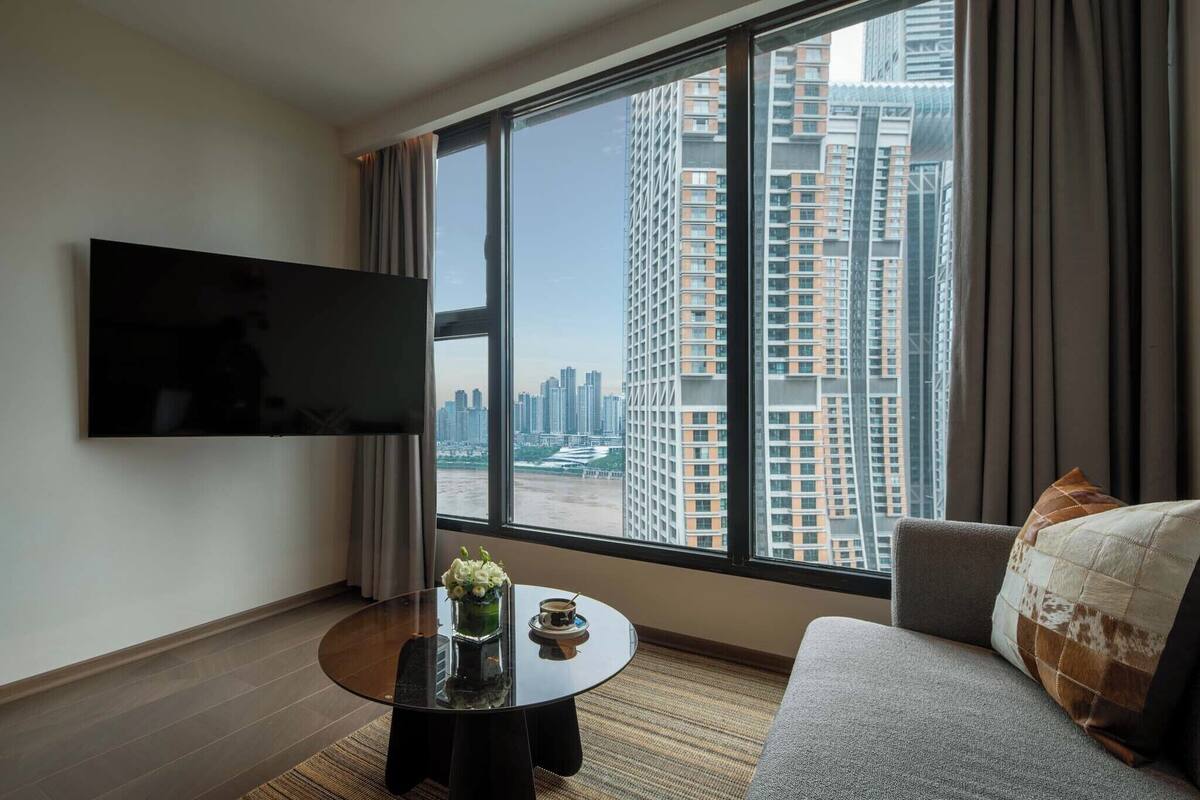 Una habitación de hotel moderna con un ventanal grande, un televisor de pantalla plana, una mesa de centro redonda con un florero y una taza, y un sofá gris con un cojín estampado.
