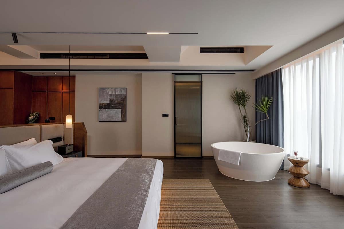 Una habitación de hotel moderna con una cama grande, una bañera, un cabecero de madera, una obra de arte colgada en la pared y un ventanal con cortinas.