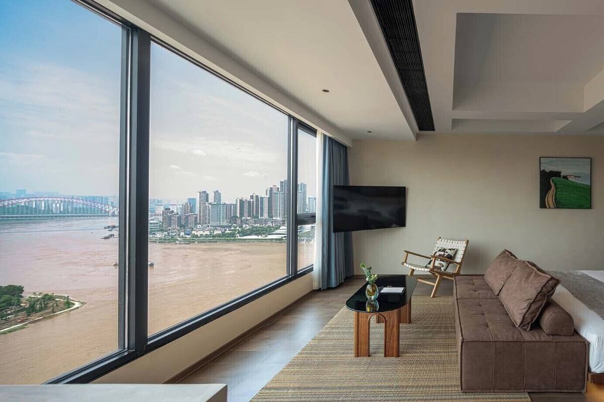 Habitación de hotel moderna con un amplio ventanal que ofrece vistas a la ciudad, un sofá cómodo, una cama y una mesita auxiliar con un jarrón.