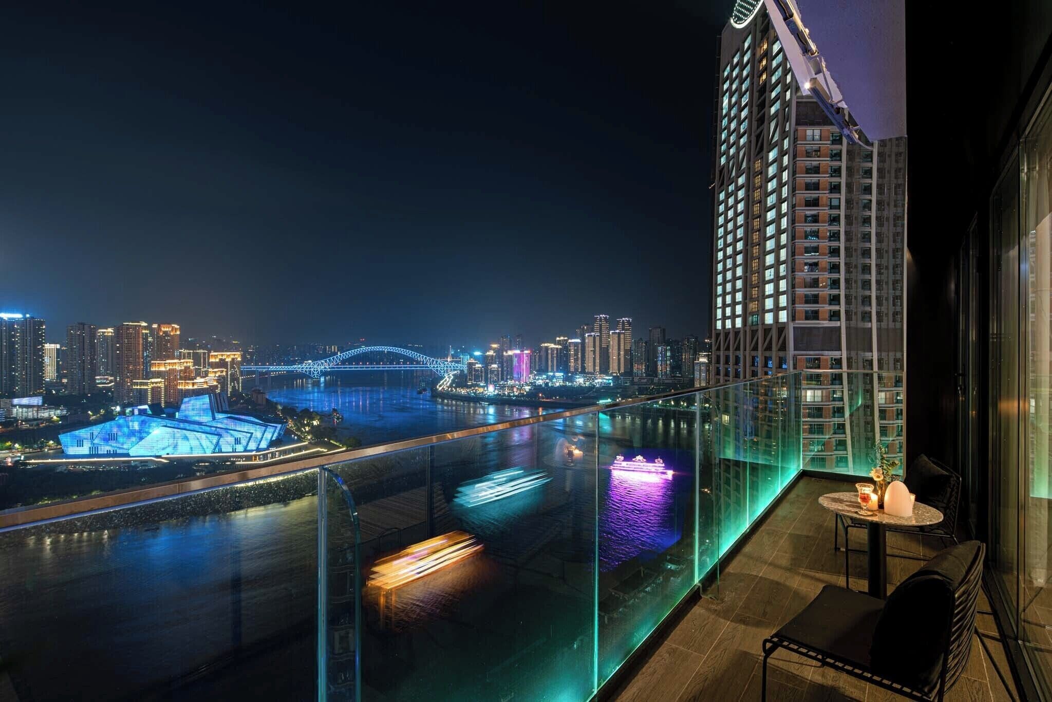 A night view of a high-rise building with a glass balcony overlooking a river and cityscape.
