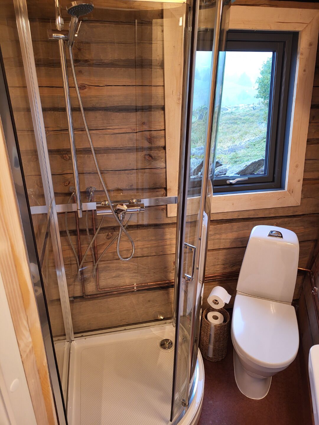 A wooden cabin bathroom with a glass-enclosed shower, a toilet, and a window with a view of the outdoors.