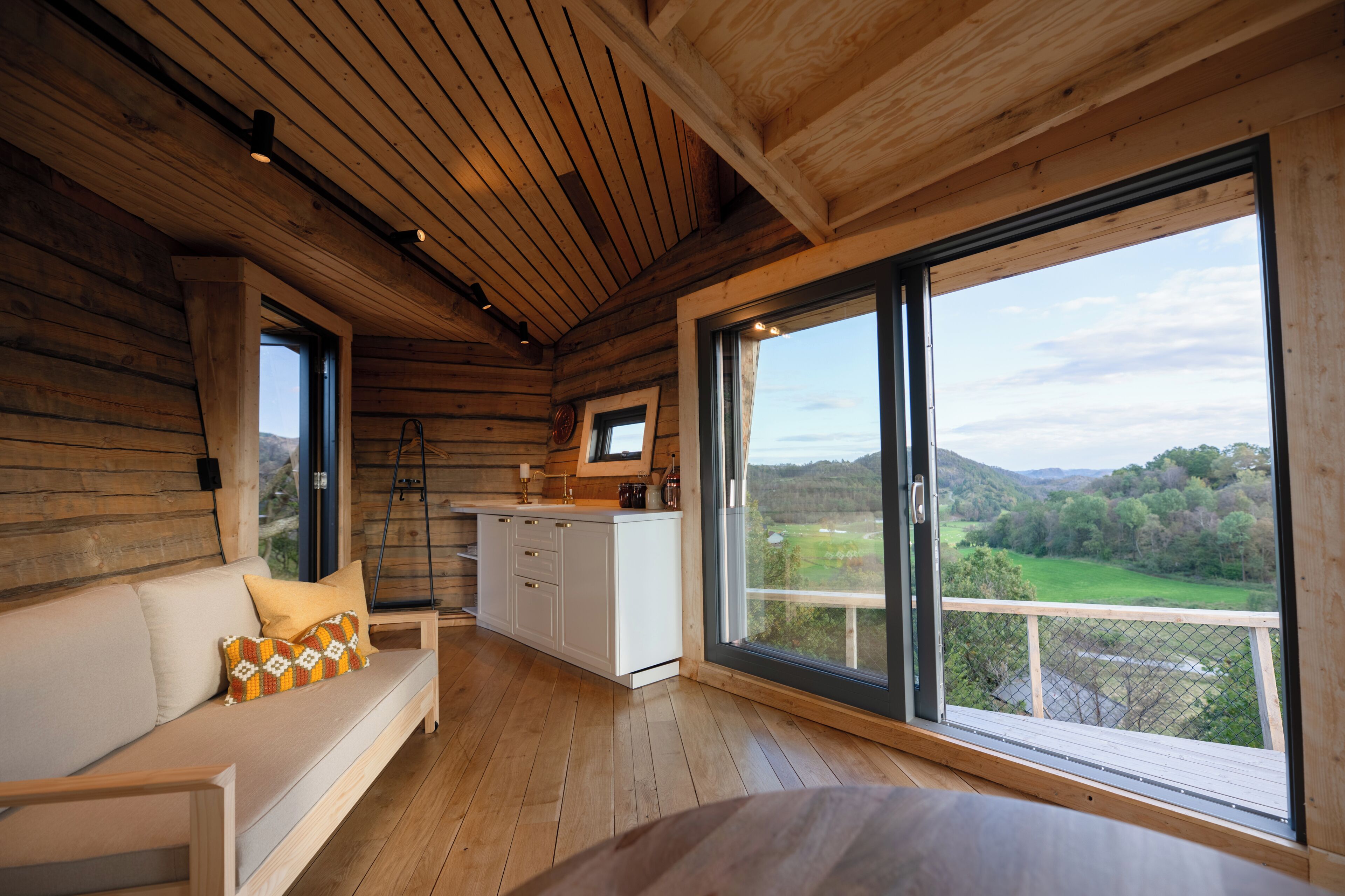 A cabin interior with a wooden floor, a sofa with cushions, a kitchen area, and a large window offering a view of the outdoors.