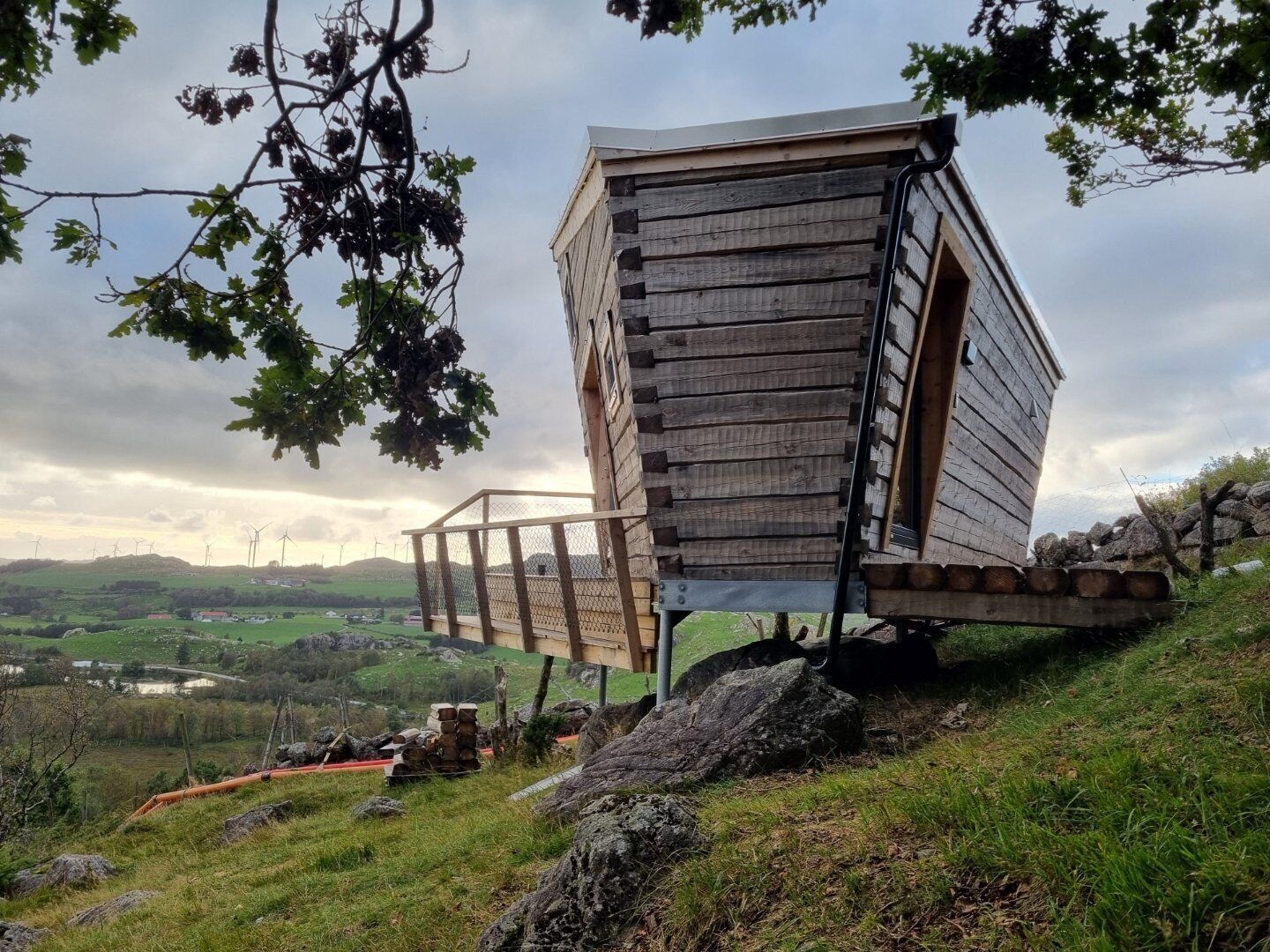 A wooden cabin with a sloped roof, situated on a grassy hill with a view of wind turbines in the distance.