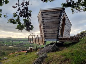 Une cabane en bois, dotée d’un toit en pente, située sur une colline herbeuse et offrant une vue sur des éoliennes au loin.