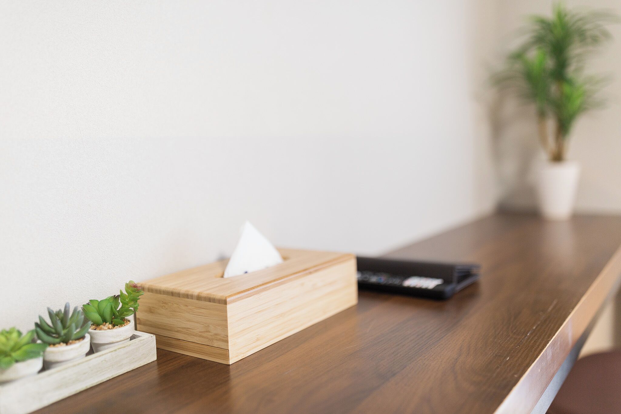 A wooden tissue box with a tissue on top, a remote control, and three small potted plants on a wooden table.