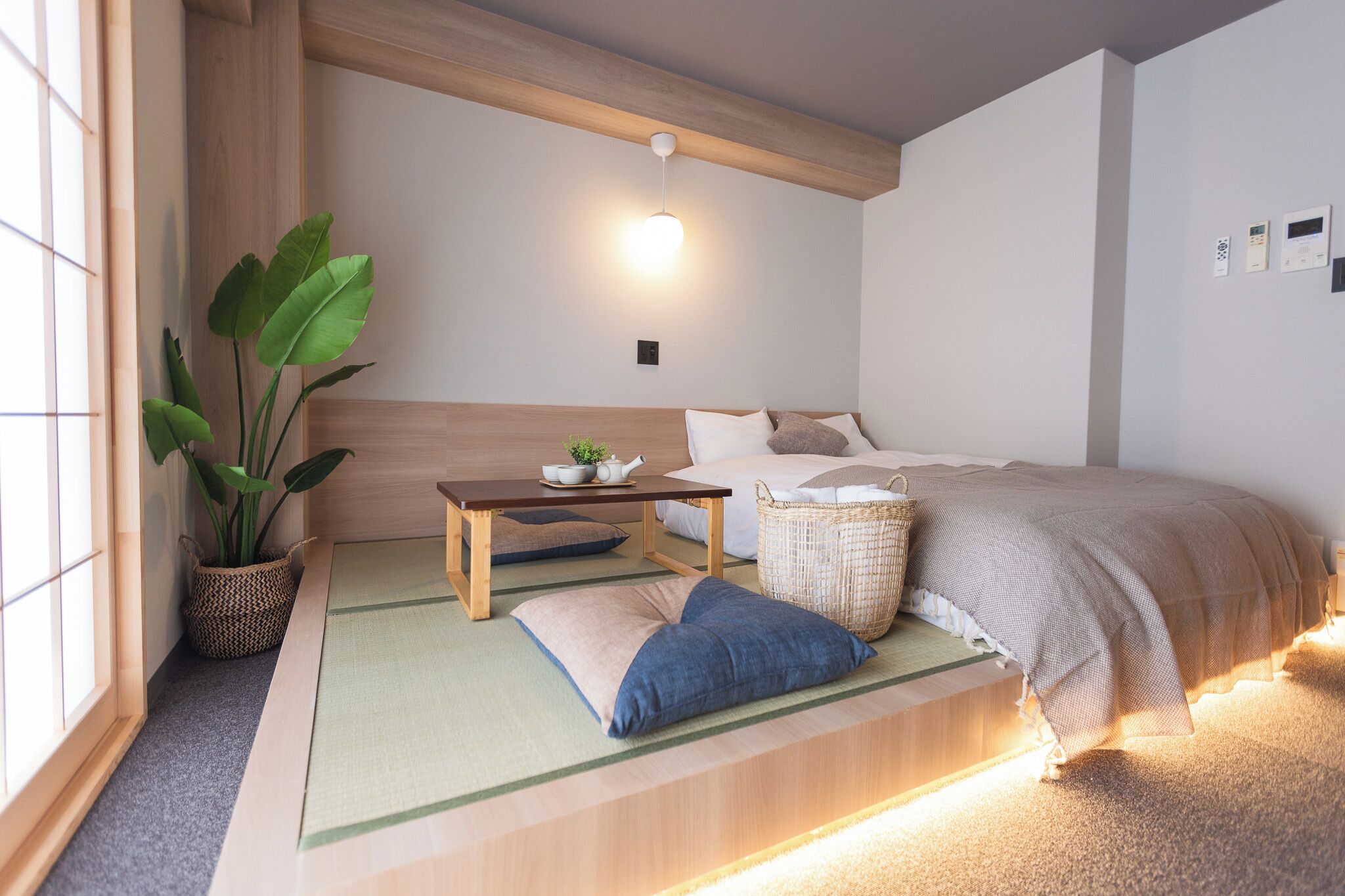 Une chambre de style japonais traditionnel, avec un sol recouvert de tatamis, un lit bas en bois, une petite table agrémentée d’une plante et un luminaire suspendu.