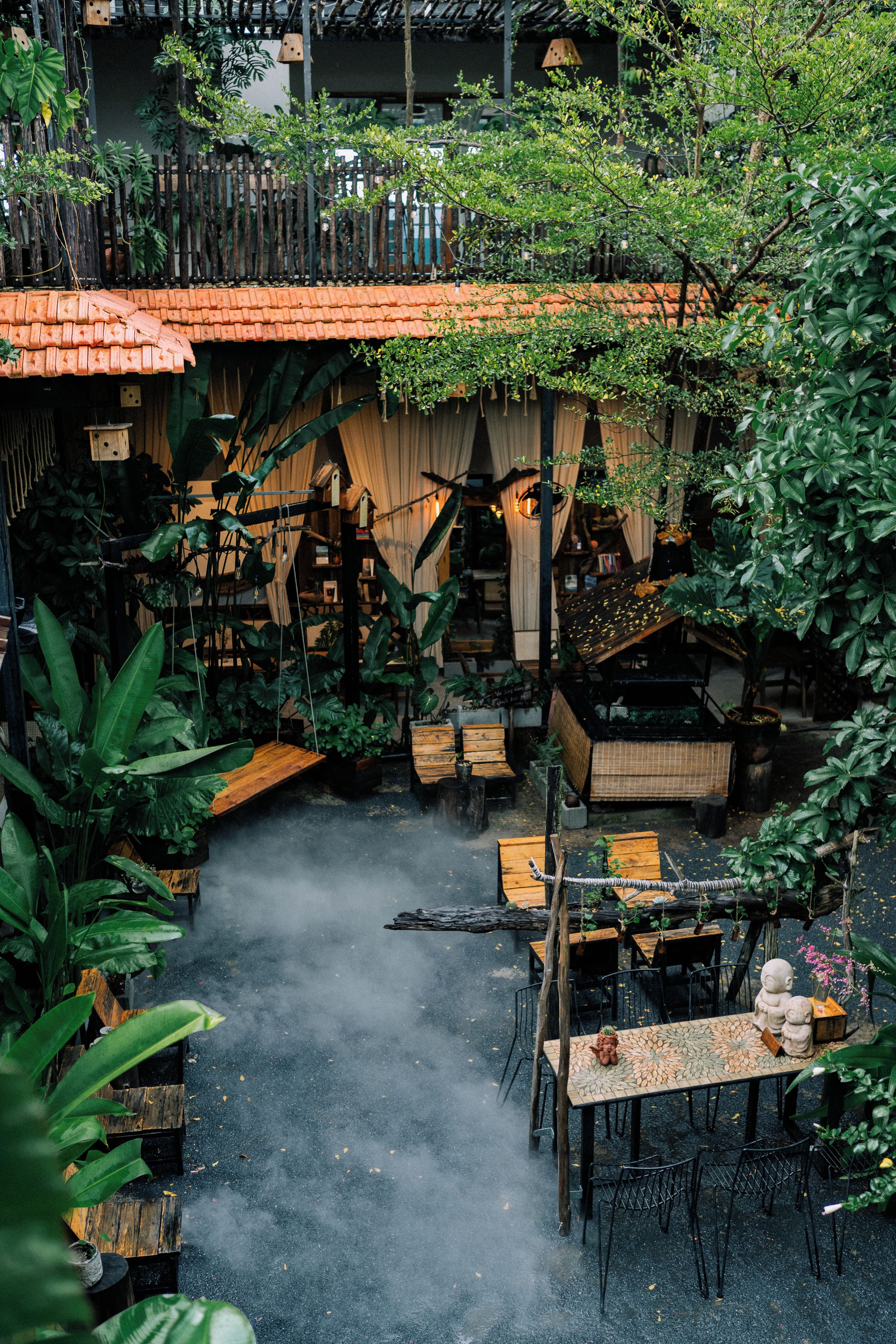 Une cour avec une table et des chaises en bois, entourée d’une végétation luxuriante et surmontée d’une structure à toit de tuiles.