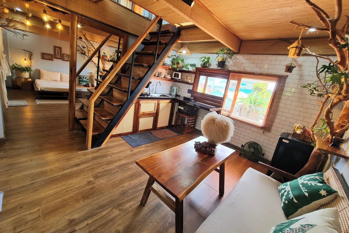 A spacious living area with a wooden staircase, a cozy sofa, a dining table, and a kitchen area with a window.