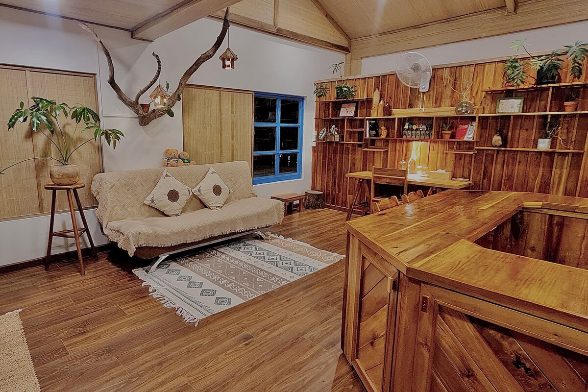 A cozy living room with a wooden floor, a sofa with cushions, a coffee table, a dining table, and a bookshelf filled with books and decorative items.