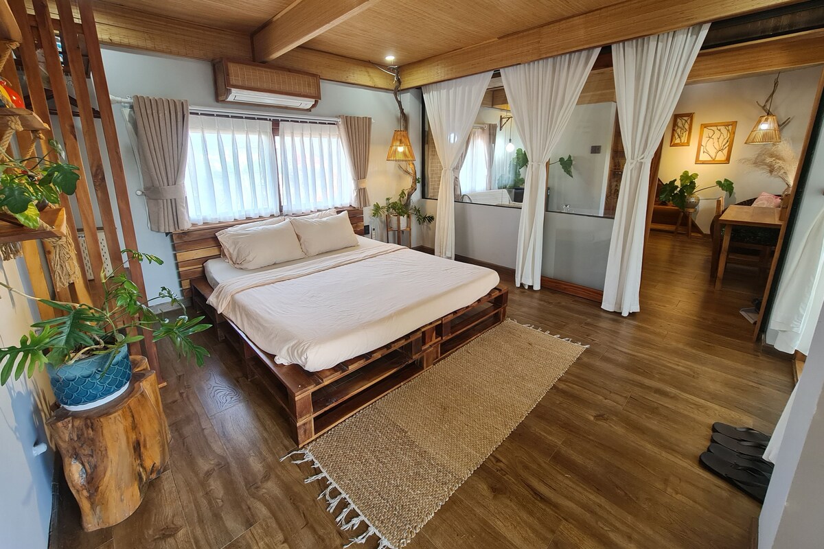 A bedroom with a wooden bed, a large window with curtains, a wooden ceiling, and a small table with a plant.