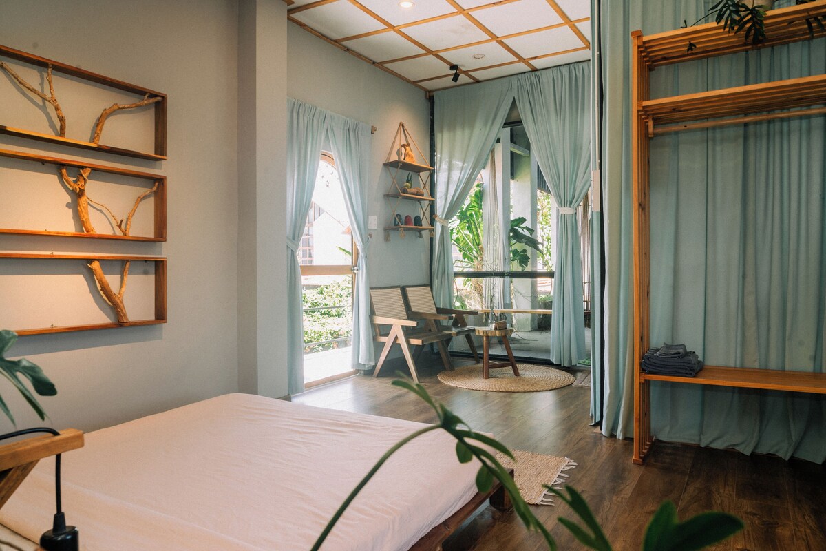 A bedroom with a bed, a desk, and a chair. There are shelves with plants and a window with curtains.