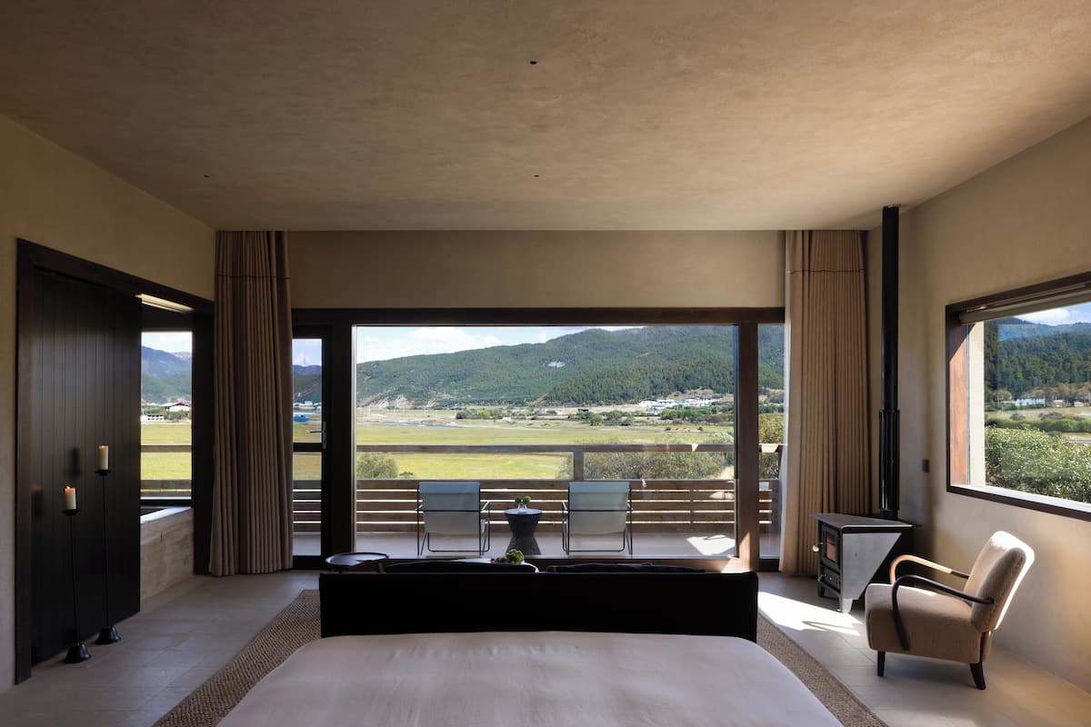 Un dormitorio con una cama grande, dos sillones, una mesita y vistas a un paisaje verde a través de un ventanal amplio.