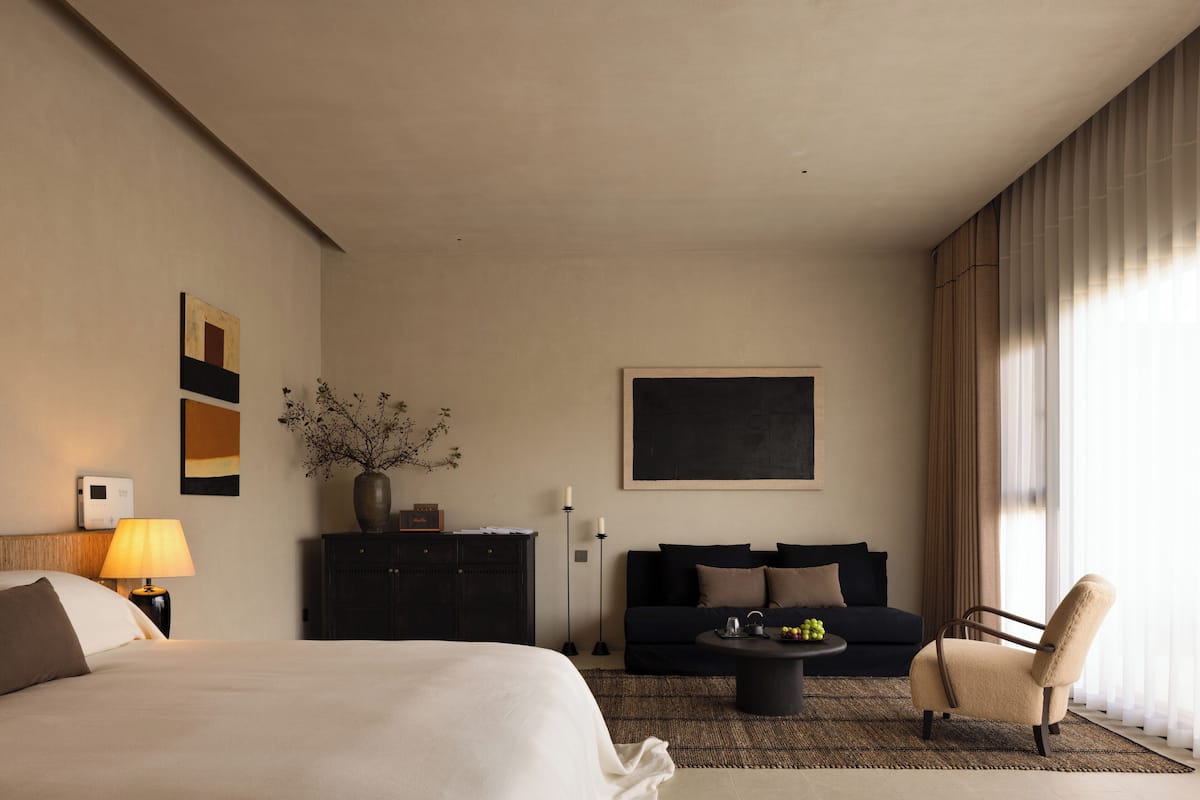 Habitación de hotel moderna con una cama grande, un sofá, una mesa de centro y una silla. Hay un cuadro en la pared y un florero con ramas sobre un mueble.