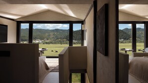 A modern hotel room with a large window offering a view of a green landscape and mountains. There is a bed, a chair, and a small table with a plant. - Shangri-La Bandu Xingye Muge Manor Hotel (Deqin)