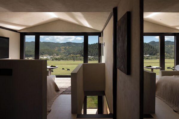 Habitación de hotel moderna con un ventanal que ofrece vistas a un paisaje verde y montañas. Se observa una cama, un sillón y una mesita con una planta.