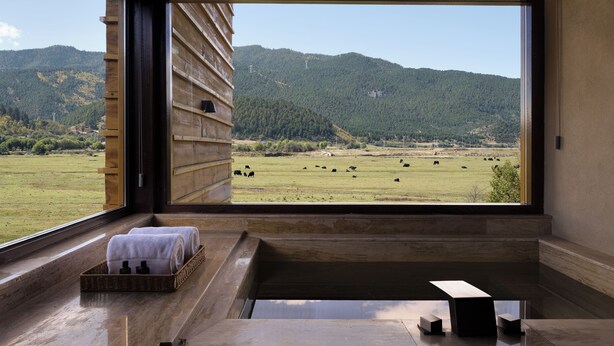 A room with a large window offering a view of a green field and mountains. There is a wooden bench and a small table with towels and toiletries. - Shangri-La Bandu Xingye Muge Manor Hotel (Deqin)