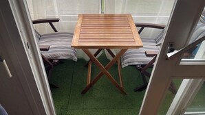 A small balcony with a wooden table and two chairs, a green artificial grass floor, and a view of the street through sheer curtains. - Le Pégase 2 (Courseulles-sur-Mer)